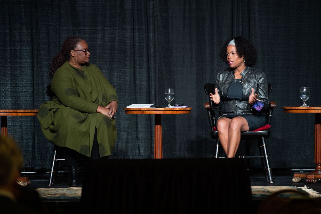Photo of a panel discussion between Deborah Douglas, Co-EIC of The Emancipator (moderating), left. She wears a forest green dress and sits with her hands on her lap, looking towards Kim Janey, Mayor of Boston, who sits to her right. Janey is wearing a gray shirt and pencil skirt and is mid-speaking. They both sit on stage with a black curtain in the background.