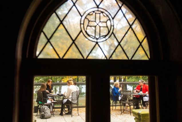 Photo taken from inside the BU Club, looking out one of the gothic, stained glass windows towards diners eating on the patio. A group of three and two are seen eating, and behind them are trees with fall-colored leaves.
