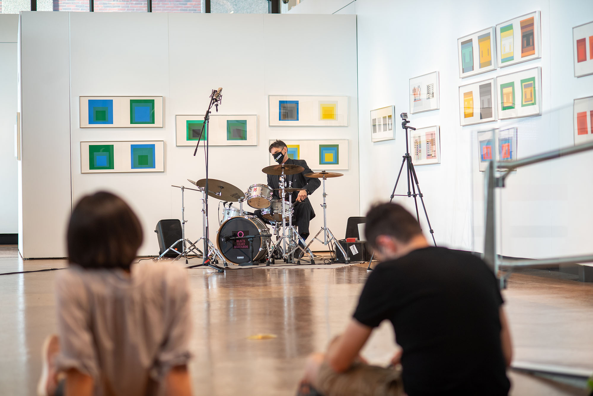 photo of Gareth Dylan Smith, Assistant Professor of Music, Music Education, playing the drums in the Stone Gallery on October 15. On the white walls behind him are colorful squares that are apart of Steve Locke’s exhibition in the gallery. Two students sit on the floor in the foreground has he plays. Gareth wears a suit and black face mask.