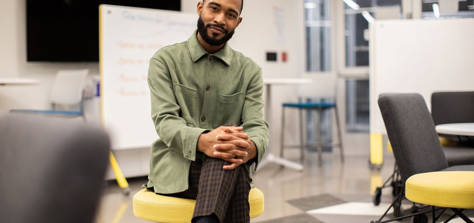 Photo of Jonathan Allen (LAW’19), a young Black man with a beard, sitting on a yellow stool. He wears a green jacket and smiles. A whiteboard is seen behind him.