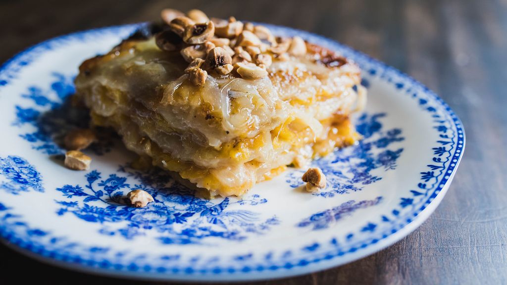 Photo of a slice of Butternut Squash and Potato Gratin topped with roasted hazelnuts on a white plate with ornate blue design. Cheese oozes from the gratin.