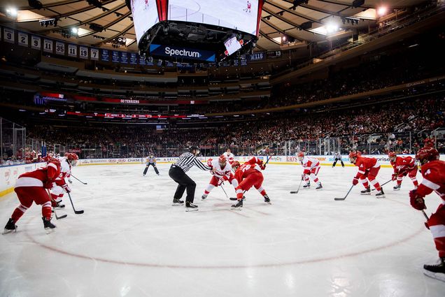 photo of the Bu men's hockey team mid-puck drop facing Cornell. Stands are crowded and both teams stand poised on the ice as a ref stands at ready.