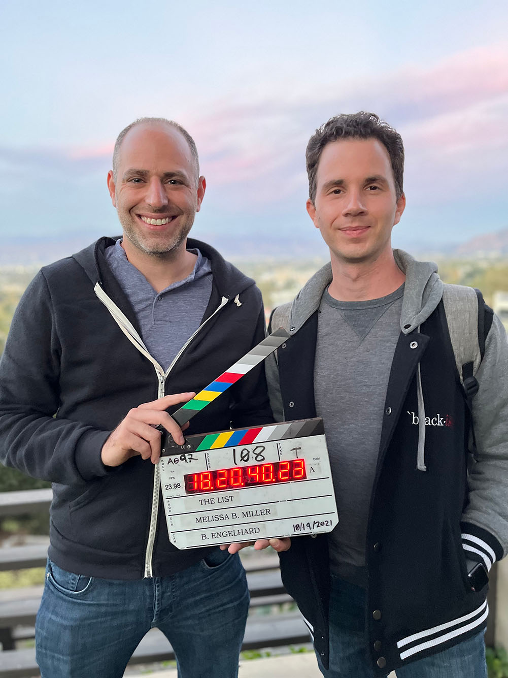 photo of Steve Vitolo and Rob Lederer on the set of their indie comedy, The List. They stand holding a clapperboard in front of a green landscape during early sunset.