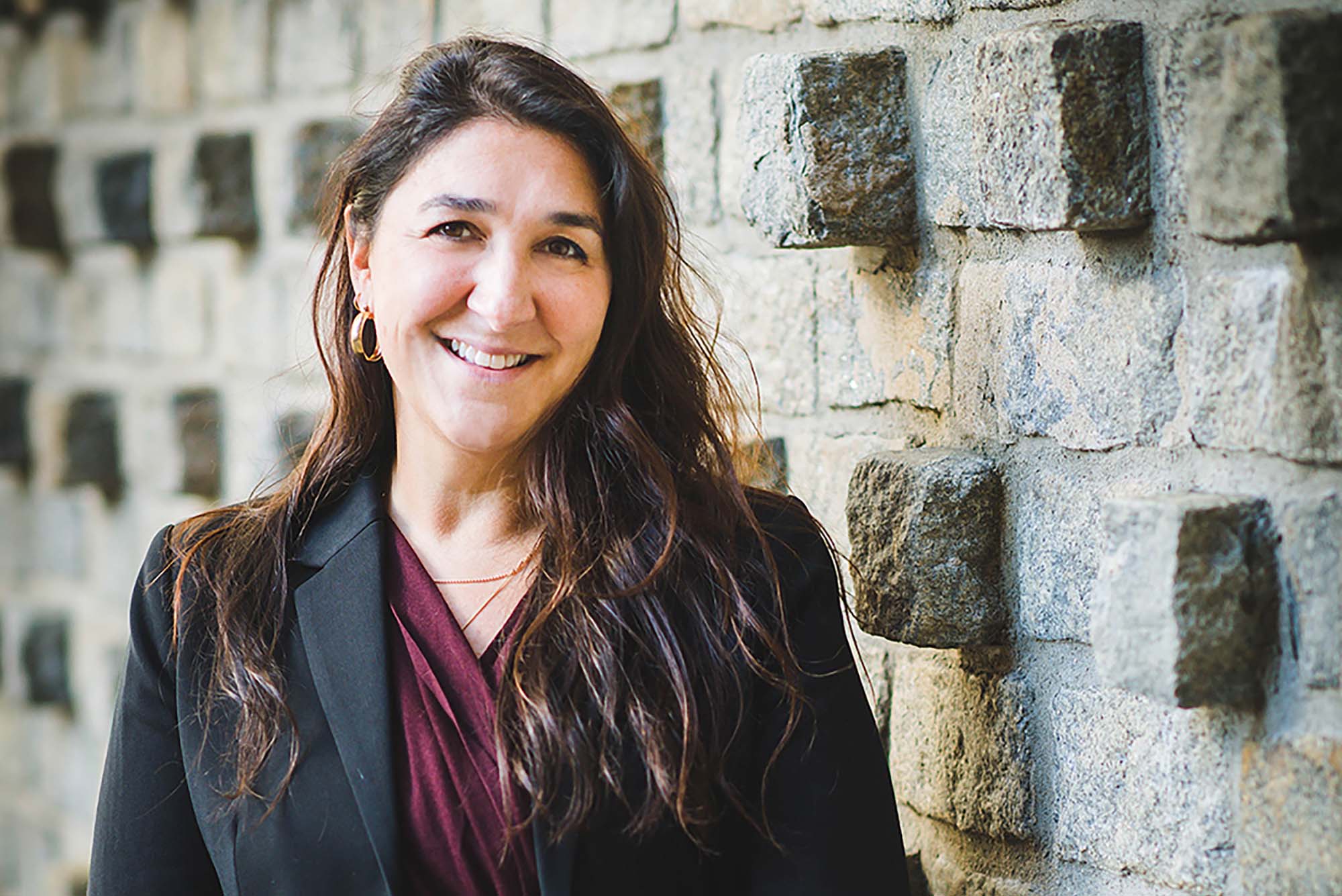 Photo of Carrie Landa, director of Behavioral Medicine and associate director of Student Health Services, is BU’s new executive director of student wellbeing. She wears a black blazer and crimson blouse and leans against a beige, brick wall.