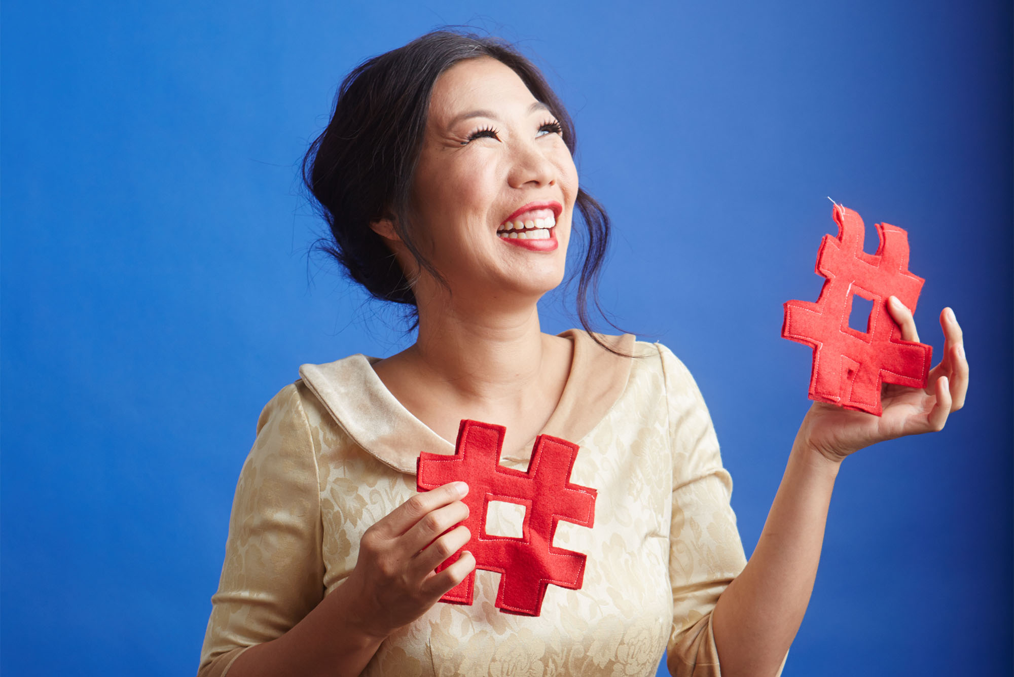 Kristina Wong stands looking upward with a smile on her face. She is holding 2 red pound symbols that appear to be made of felt.