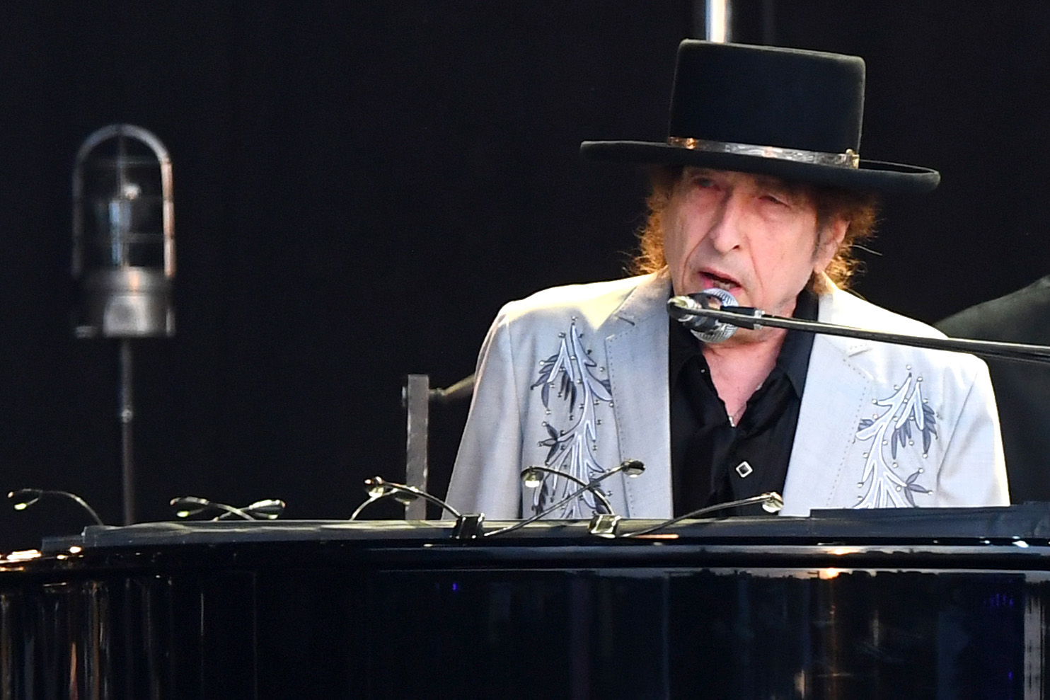 photo of Bob Dylan performing in London in 2019. He wears a black hat and light jean jacket as he sings into a microphone as he plays a black piano on stage.