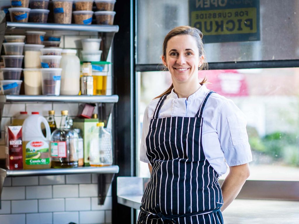 Photo of BU alum Kate Smith, who is the co-head owner and chef at Boston-area gastropub Thistle & Leek. She wears a white short sleeved chef's jacket with a blue, white-striped apron over it. Behind her, small prep containers of food are seen.