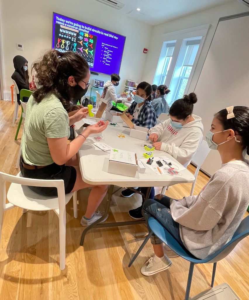 Photo of Michalina (Mica) Jadick (green shirt at left), one of the BU STEM undergraduates who worked part of the team with Paul Trunfio on co-designing the Network Science curriculum. In the photo she is supporting the youth in her group to create their own circuits to light the light bulb. Mica sits at a low, white table with a group of young people as they complete an activity. Everyone wears face masks.