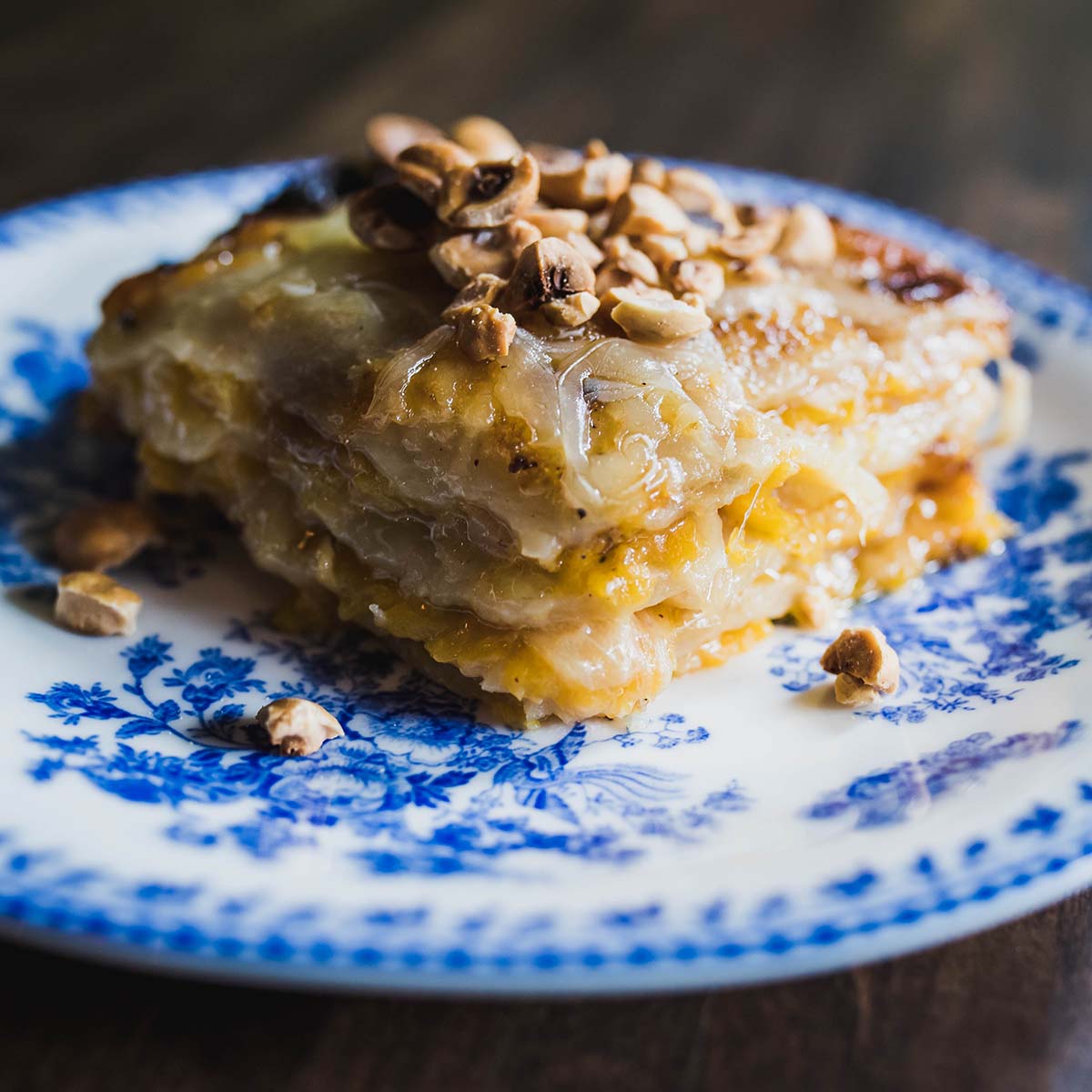 Photo of a slice of Butternut Squash and Potato Gratin topped with roasted hazelnuts on a white plate with ornate blue design. Cheese oozes from the gratin.