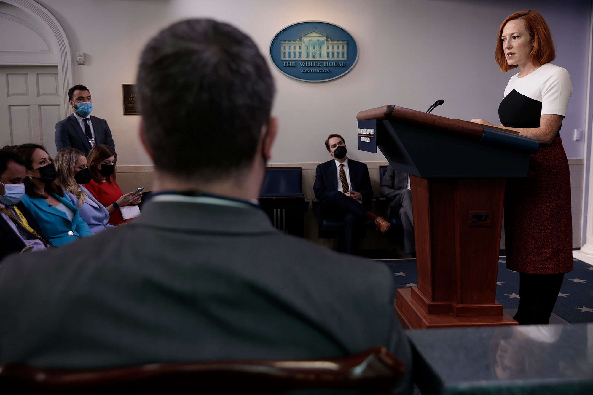 photo of White House Press Secretary Jen Psaki talks to reporters during the daily press conference in the Brady Press Briefing Room at the White House on December 06, 2021 in Washington, DC. She wears a black and white dress as she stands at a lectern and speaks to seated reporters.