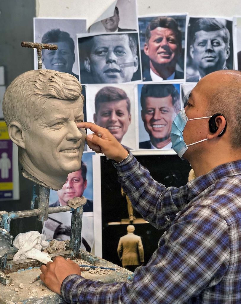A scultptor sits in front of a photoboard filled with pictures of JFK, as he models JFK's likeness out of clay