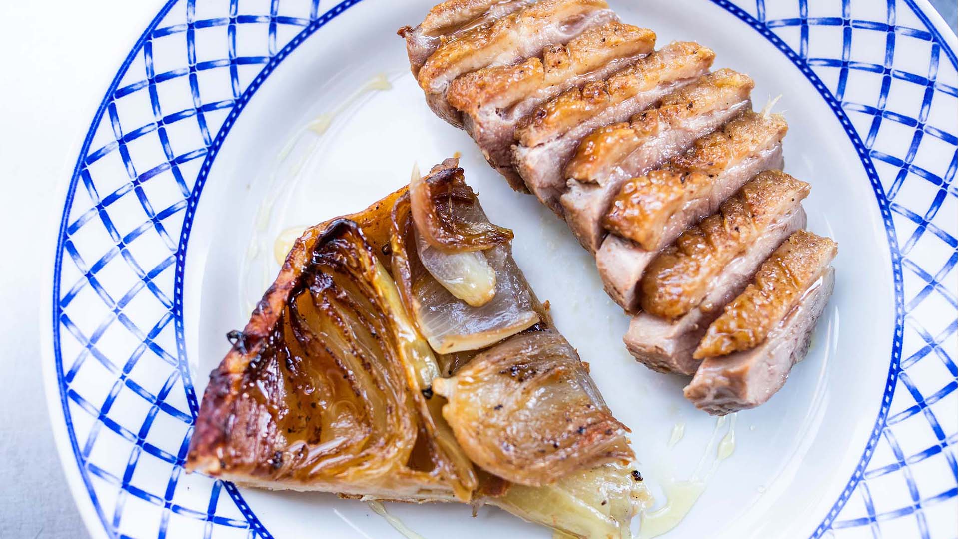 Photo of a sliced Duck Breast and Onion Tarte Tatin on a white plate with a blue border.