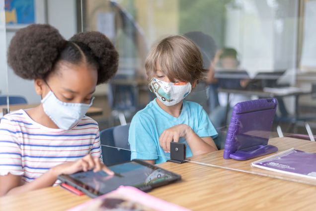 two school children sit next to each other with a plastic divider between them and look at a tablet together