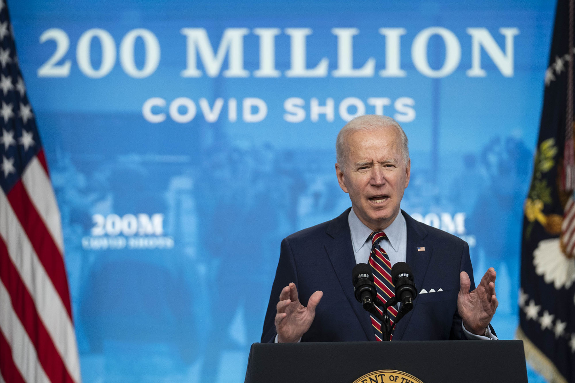 Photo of Biden addressing his administration’s vaccine efforts while speaking in the Eisenhower Executive Office Building in Washington, D.C., last year. His hands are raised mid-gesture as he speaks at a podium.