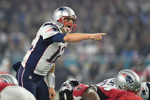 Photo of Tom Brady playing during Super Bowl LI at the NRG Stadium in Houston, TX, USA. He points forward and yells in his Patriots uniform as his teammates and the opposing team are in "hut" stance around him.