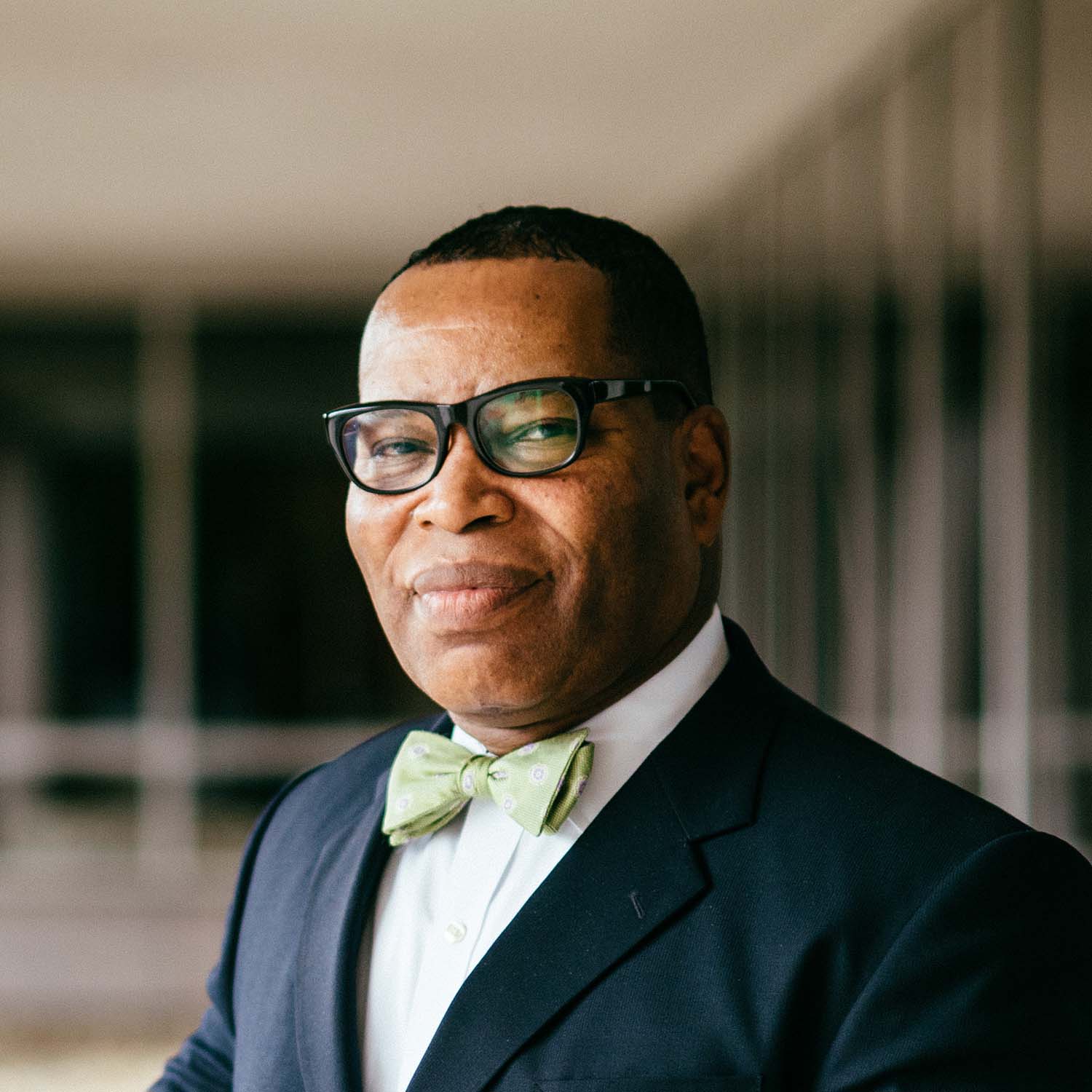 Photo of Ralph Groce, a middle-aged Black man with thick black glasses wearing a blue suit and green bow tie as he leans against a railing and clasps his hans.
