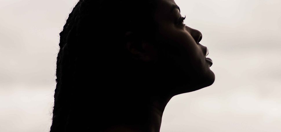 Photo of the side profile of a dark-skinned black woman, bare from the shoulder and with long braids, looks off to the right. The profile is mostly in shadow, though the tones of her skin are noticeable. Background is a hazy white, similar to clouds.