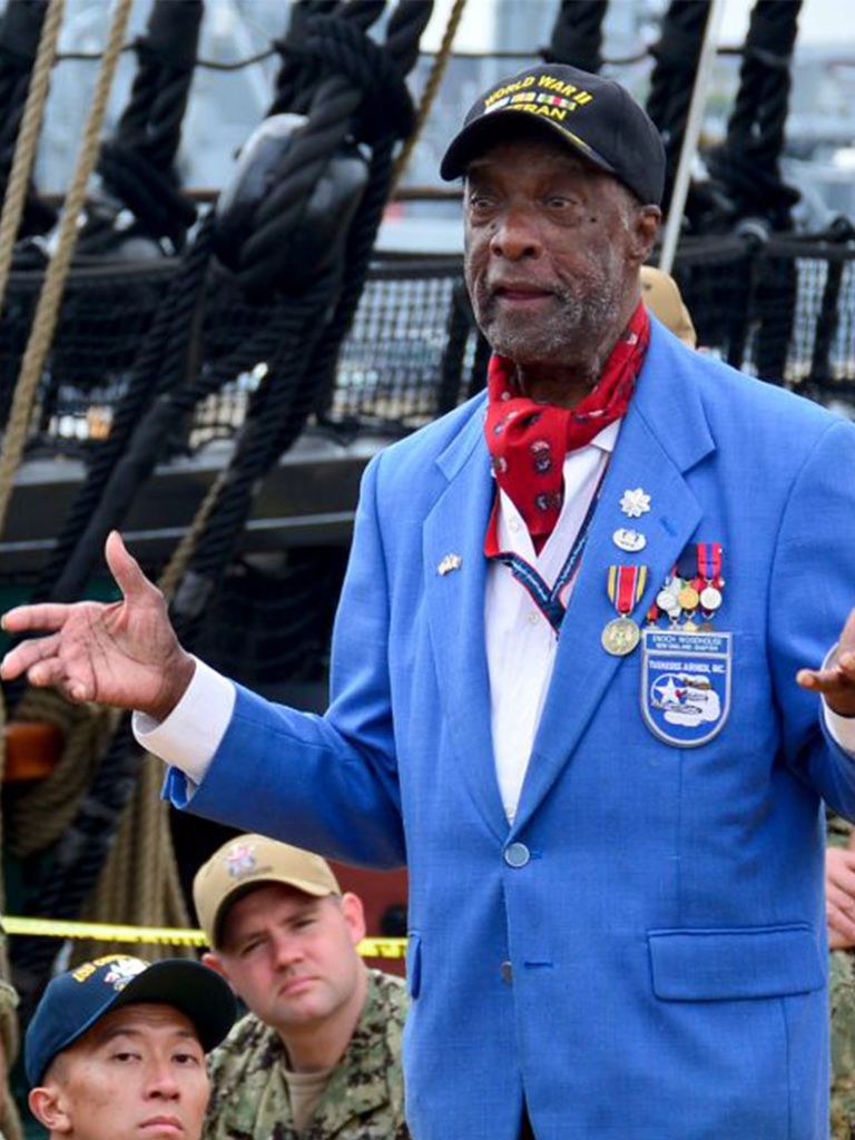 Photo of Retired U.S. Air Force Lt. Col. Enoch Woodhouse in a blue suit jacket, baseball cap, and red neck-tie with his hands extended towards a crowd of chief petty officer selects aboard USS Constitution, who wear camo and tan baseball caps. US Navy photo by Mass Communication Specialist 3rd Casey Scoular/Released