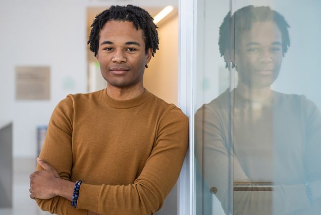 Photo of Ethan Strauther, a young Black man with short dreads wearing a burnt yellow sweater. He leans against a glass wall that reflects his image back.