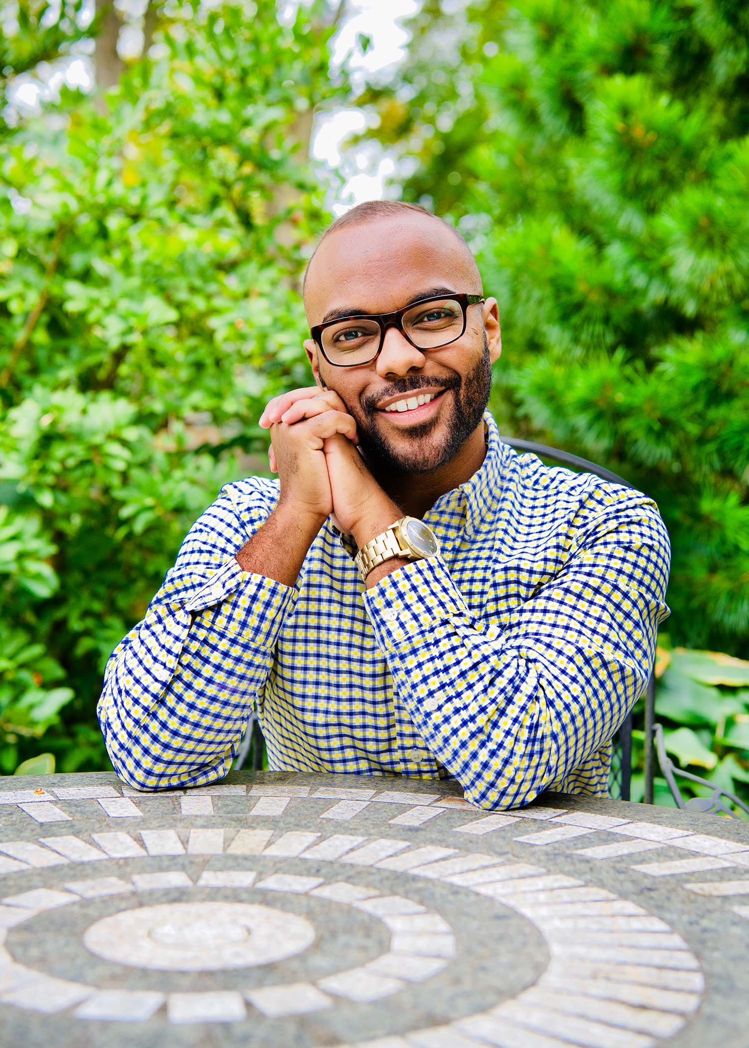 Photo of Thomas Andrews in a white and blue patterned button-down shirt with his hands clasped. He is a young man of color with black-rimmed glasses, smiling, as he sits at a patio table.