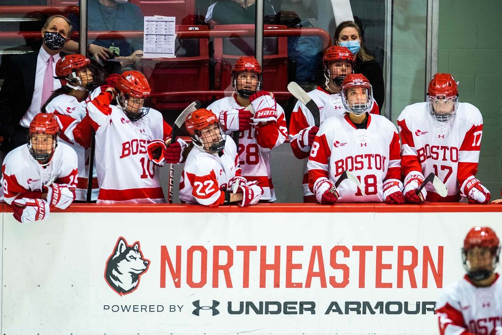 Photo of the BU women's hockey team at their bench looking on as Harvard scores a goal in the third quarter during the women’s Beanpot Tournament at Matthews Arena on February 1, 2022. Women stand with red helmets on and sticks in hand as they watch.
