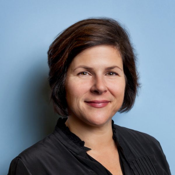 Photo of Kabrina Chang , a white woman with short brown hair, who wears a black blazer, and poses in front of a blue background.