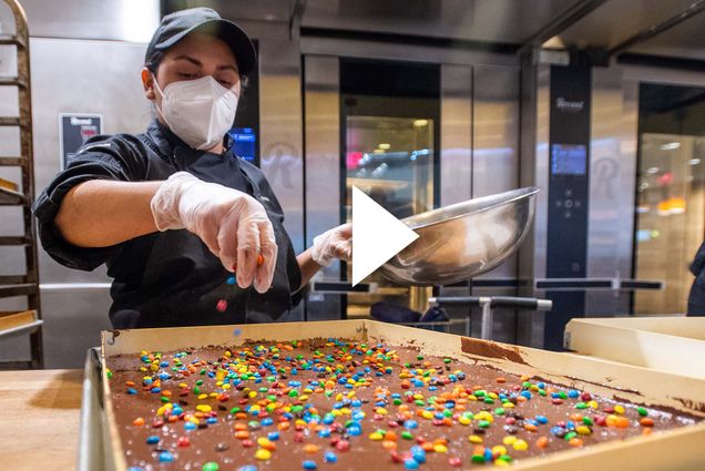 Carmen Quijada adds sprinkles to a batch of brownies. There is a white play button overlayed on top of the image