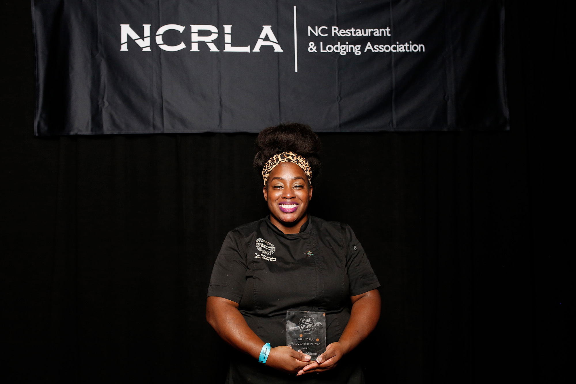 Photo of Pastry chef Tie Whittaker (MET’12), owner of dessert company Buttermilk Boutique, holding an award she won in front of a black background with white text that reads the "NC Restaurant and Lodging Association." Whittaker is a young Black woman, wearing a black chef's jacket and cheetah print headband, smiles as she holds the glass award in her hands.