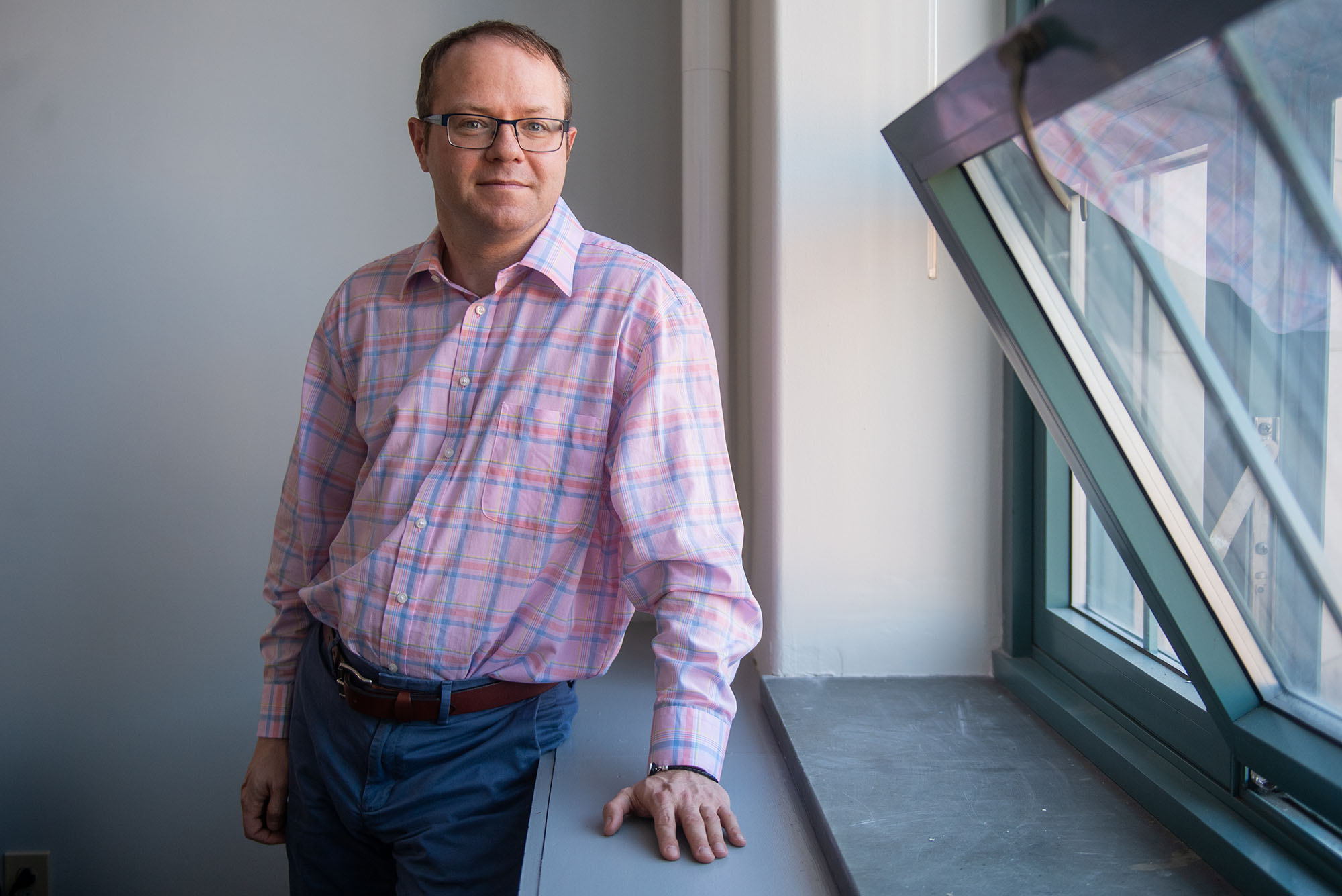 Photo of Benjamin Sovacool. A middle-aged white man wearing glasses and a light pink plaid shirt and jeans poses next to an open window and lightly smiles at the camera.