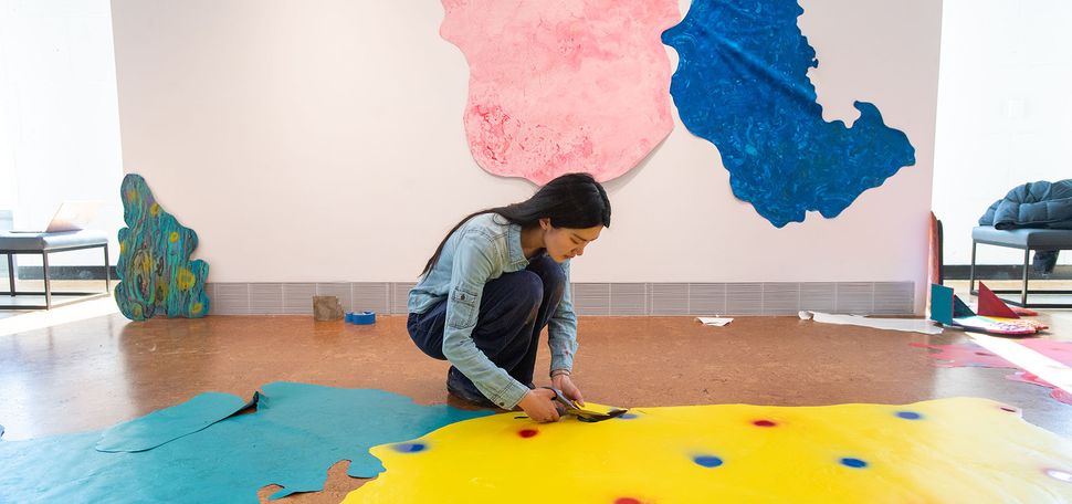 Photo of Moxin “Sunny” Chen (CFA’22) installing her vinyl-and-paint collage piece, “Yesterday Is Passed, Tomorrow Has Yet to Come". A young, Asian woman bends down and adjusts a large yellow blob that features near many other various colored blobs in the exhibit.