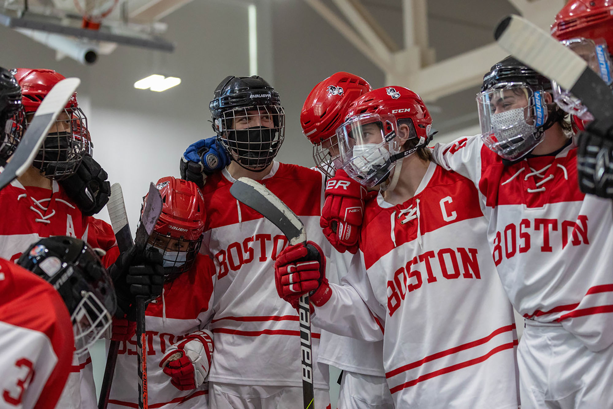 Photo of the BU Roller Hockey team in a huddle. They wear red and white jerseys that read "Boston", red helmets, and hold sticks as they huddle together.