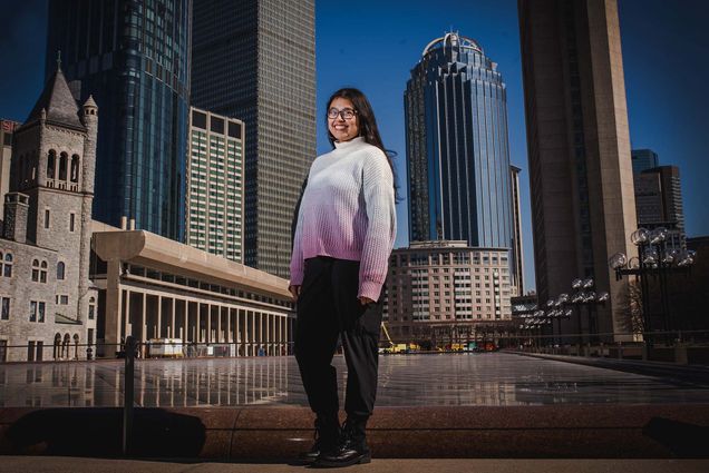 Photo of student Mira Dhakal, standing at the large reflection pool near the Christian Science Center. She smiles, wears a white and pink gradient sweater, and has long brown hair and glasses. The Boston city skyline and blue sky are seen behind her.