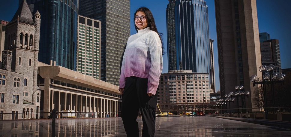 Photo of student Mira Dhakal, standing at the large reflection pool near the Christian Science Center. She smiles, wears a white and pink gradient sweater, and has long brown hair and glasses. The Boston city skyline and blue sky are seen behind her.