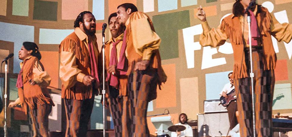 Still from the movie Summer of Soul featuring Fifth Dimension during a performance. They wear orange, yellow and brown outfits, including jackets with fringe and bell bottom pants. The women at right at the microphone raises her hands as she sings. Three men at right sing into one microphone.