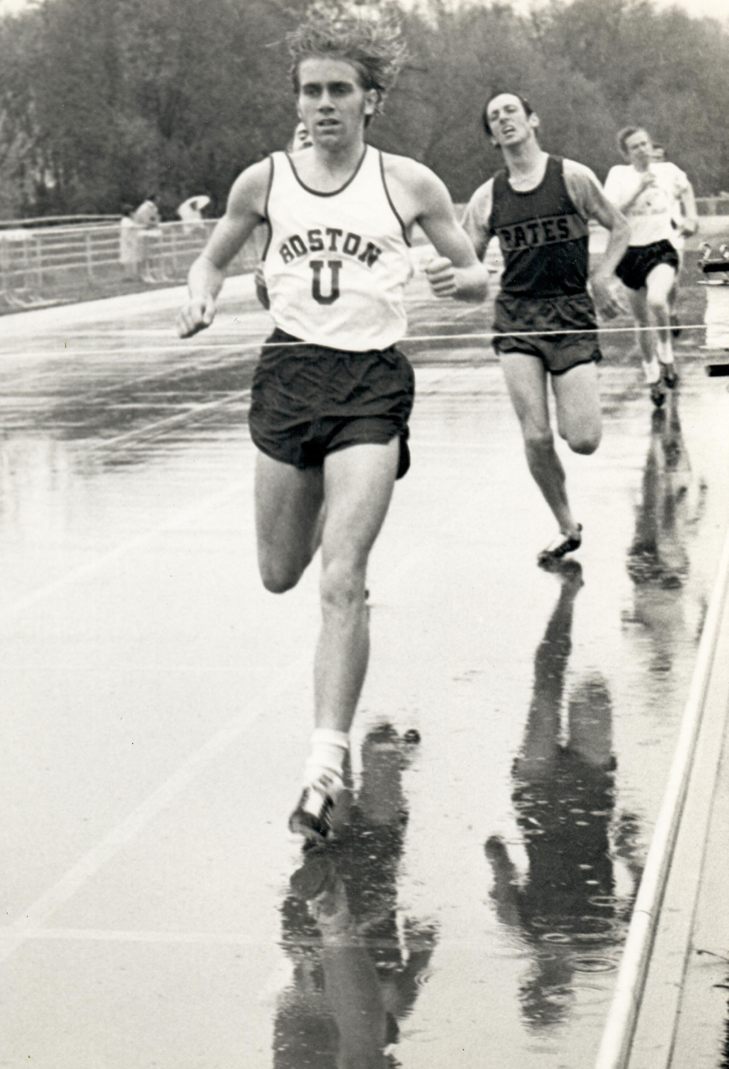 Black and white photo of Tom Beatty (CAS’72) during a historic track meet. He wears a BU jersey and his hair bounces as he runs ahead of two competitors on a track.