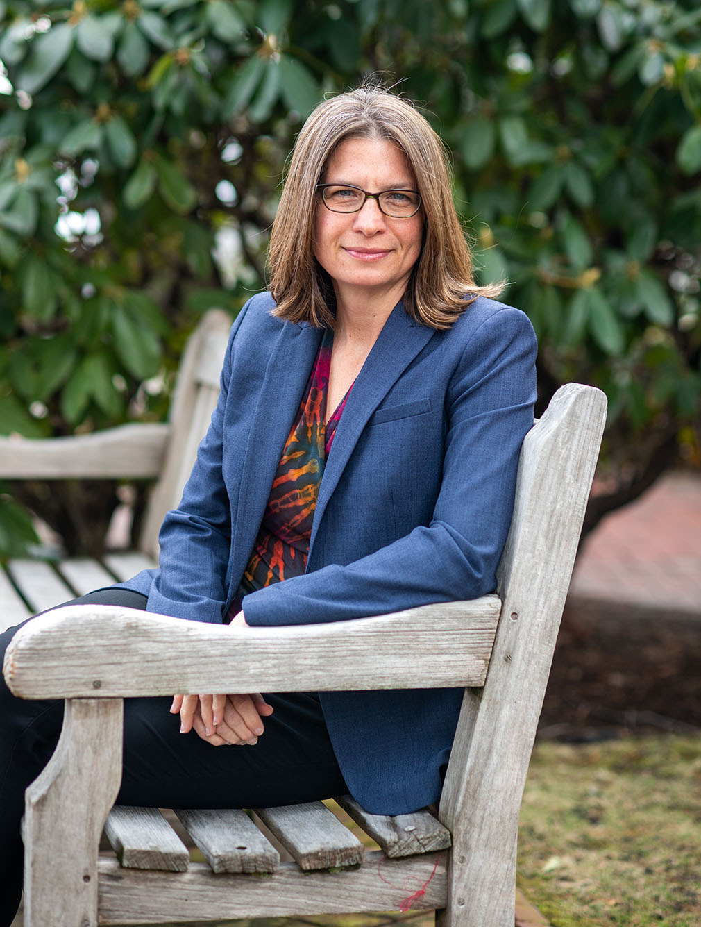 Photo of Brenda Philips sitting on a wooden bench in a park and turned to look at the camera. A white woman with glasses wearing a blue blazer, red, green, and yellow blouse, and black pants, poses for the camera.