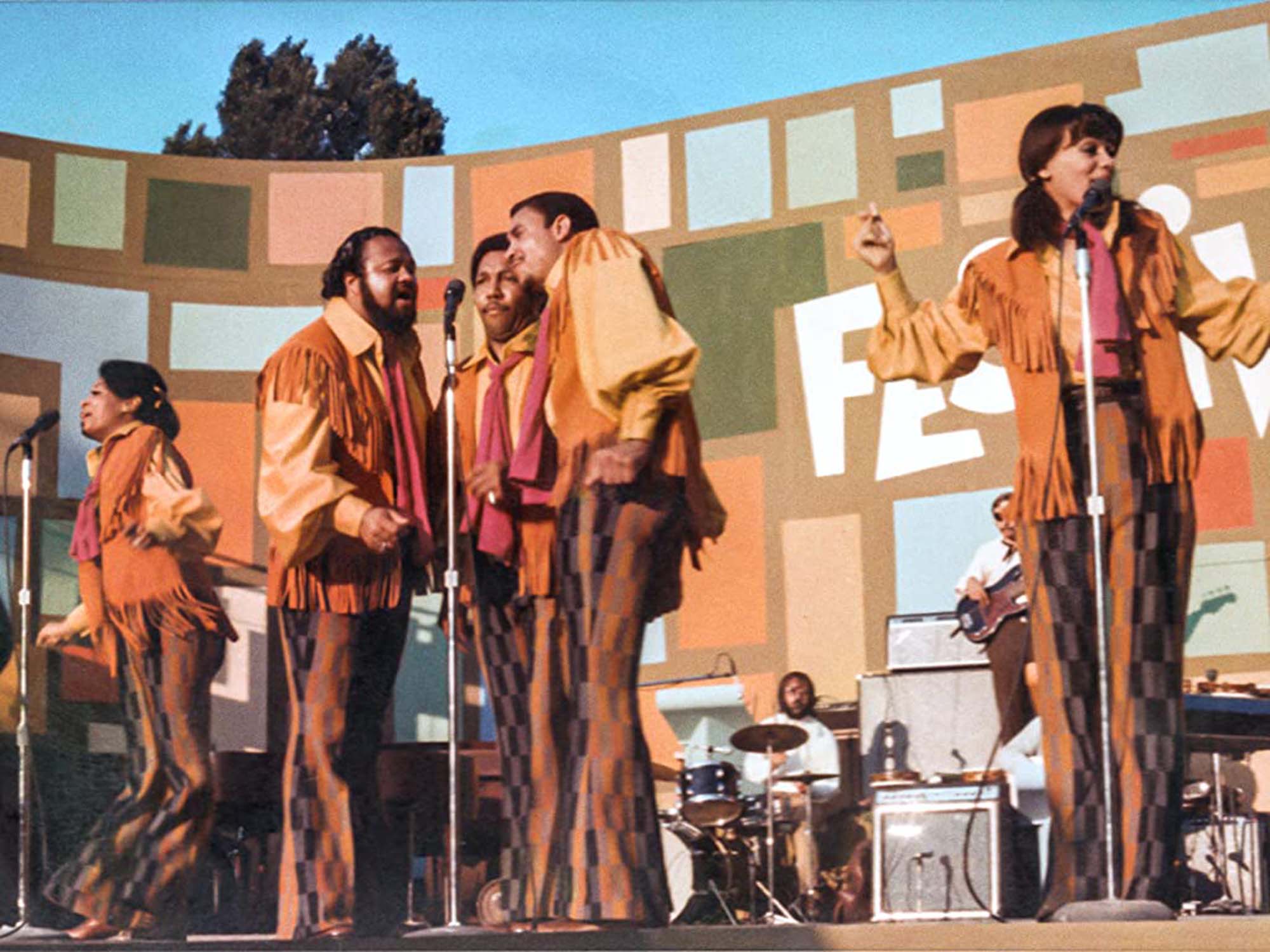 Still from the movie Summer of Soul featuring Fifth Dimension during a performance. They wear orange, yellow and brown outfits, including jackets with fringe and bell bottom pants. The women at right at the microphone raises her hands as she sings. Three men at right sing into one microphone.