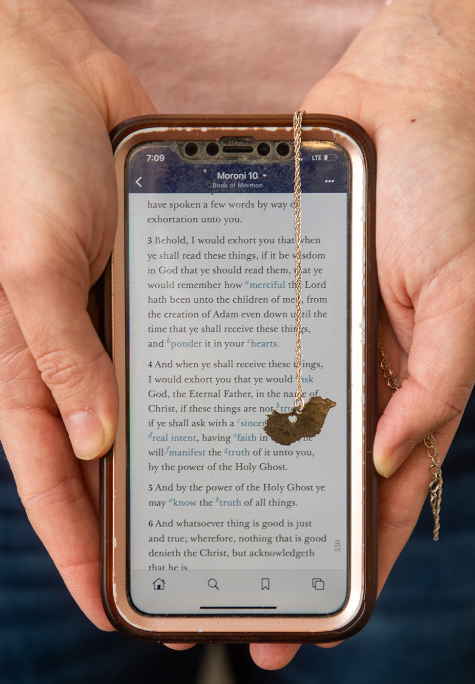 Photo of a phone being held face up in someone's hands. The screen displays a scripture from the Book of Mormon