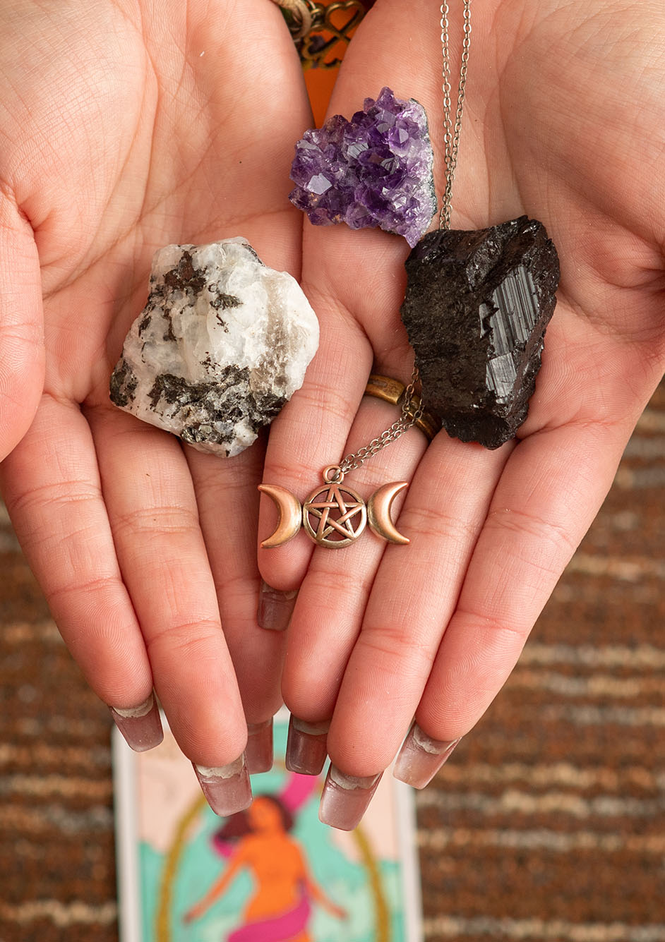 Photo of hands palm up showing three crystals and a necklace to the camera. From left, moonstone, purple amethyst, and black tourmaline. Pendants show a triple moon symbol and a pentagram.