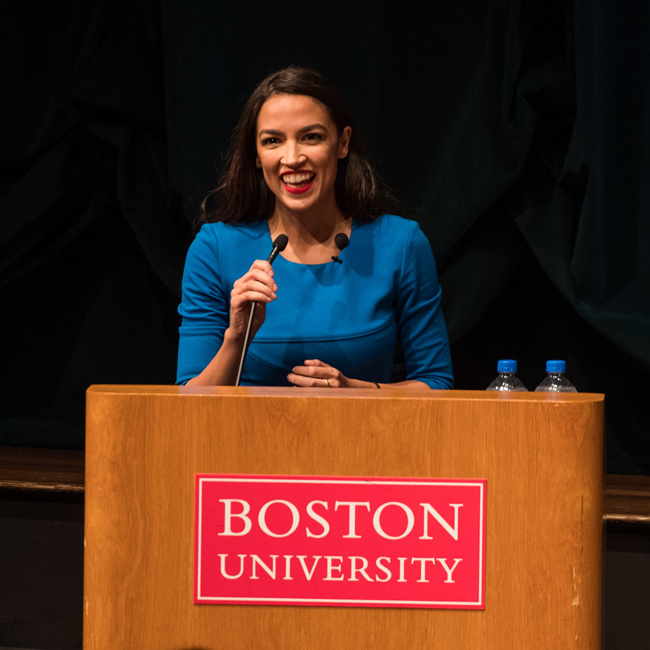 Candid photo of Alexandria Ocasio-Cortez on stage at Tsai Center. A hispanic woman wearing a blue dress is mid-laugh as she stands behind a wooden podium with a sign on front that reads "Boston University"