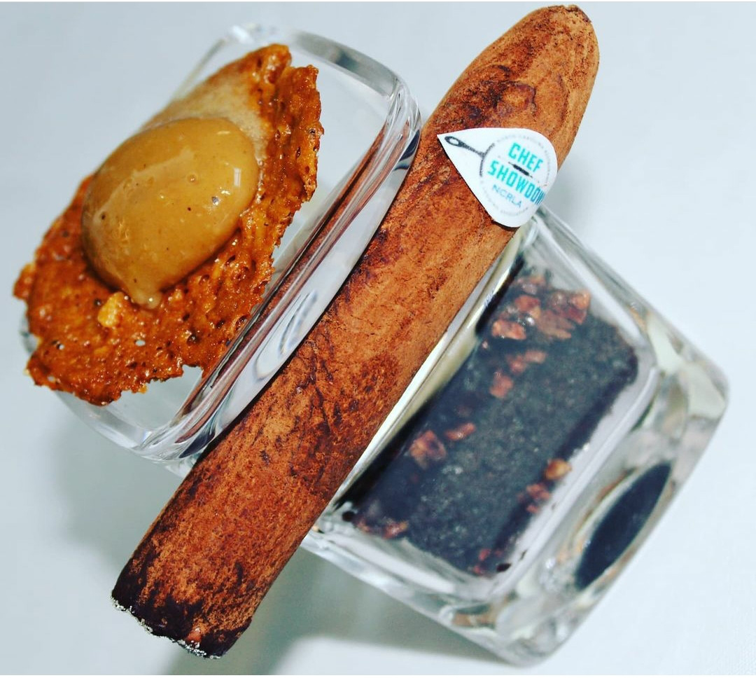 Photo of The Aficionado, Whittaker’s winning chocolate cigar dessert. Photo shows a small shot glass filled with a chocolate cake with intricate gold decoration as a delicate cigar-shaped chocolate is placed gently next to it