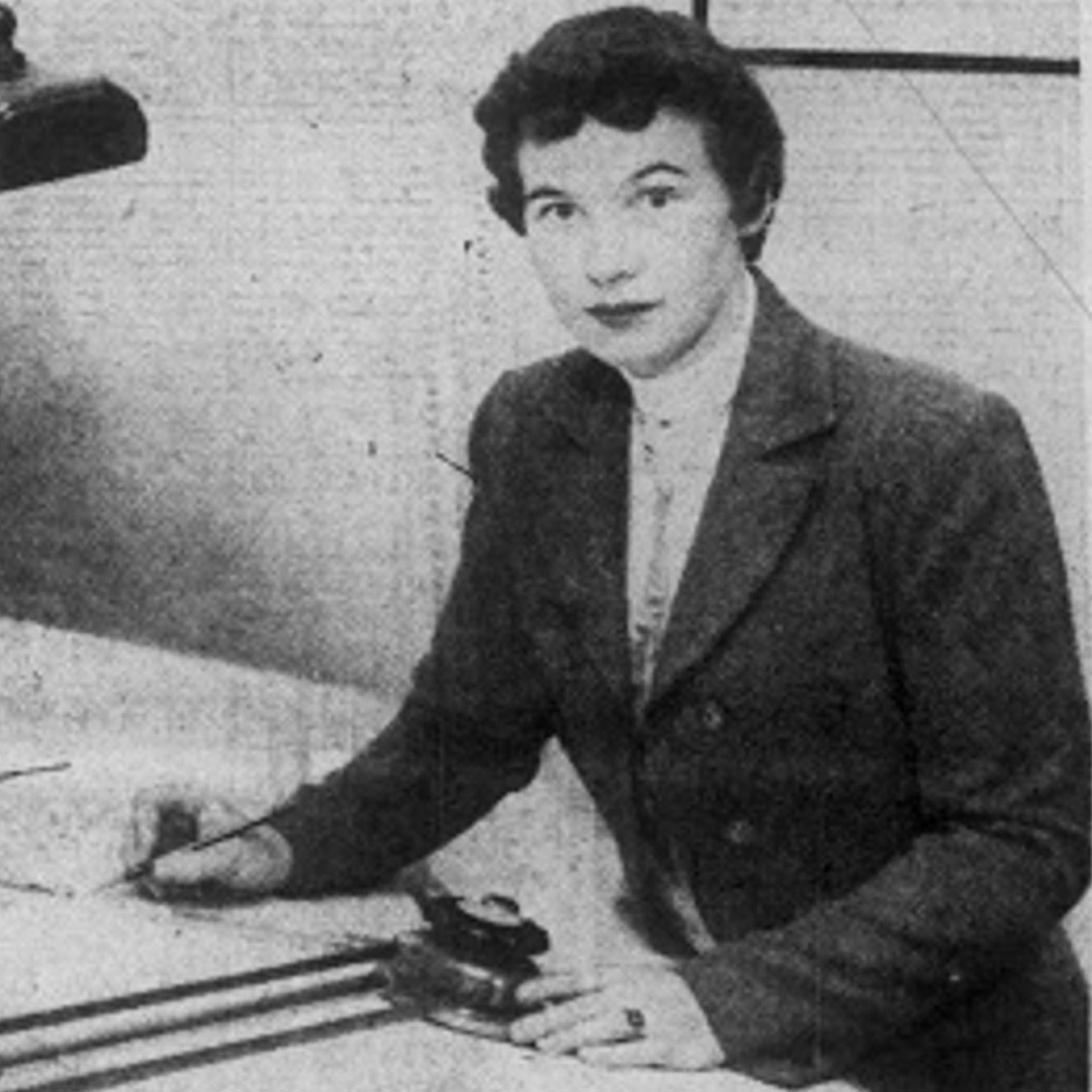 Photo of Anne Everest Wojtkowski (1935-2014), the first woman to graduate from the Boston University College of Industrial Technology. The photo is black and white and pictures Wojtkowski writing at a desk with a lamp. She has short dark hair.
