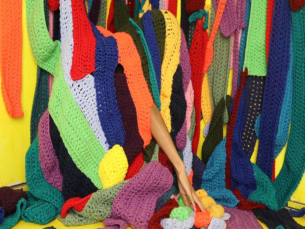 Image: Photo of a large curtain of knitted patterns of various colors sewn together. A hand can be seen reaching out from the large fabric.