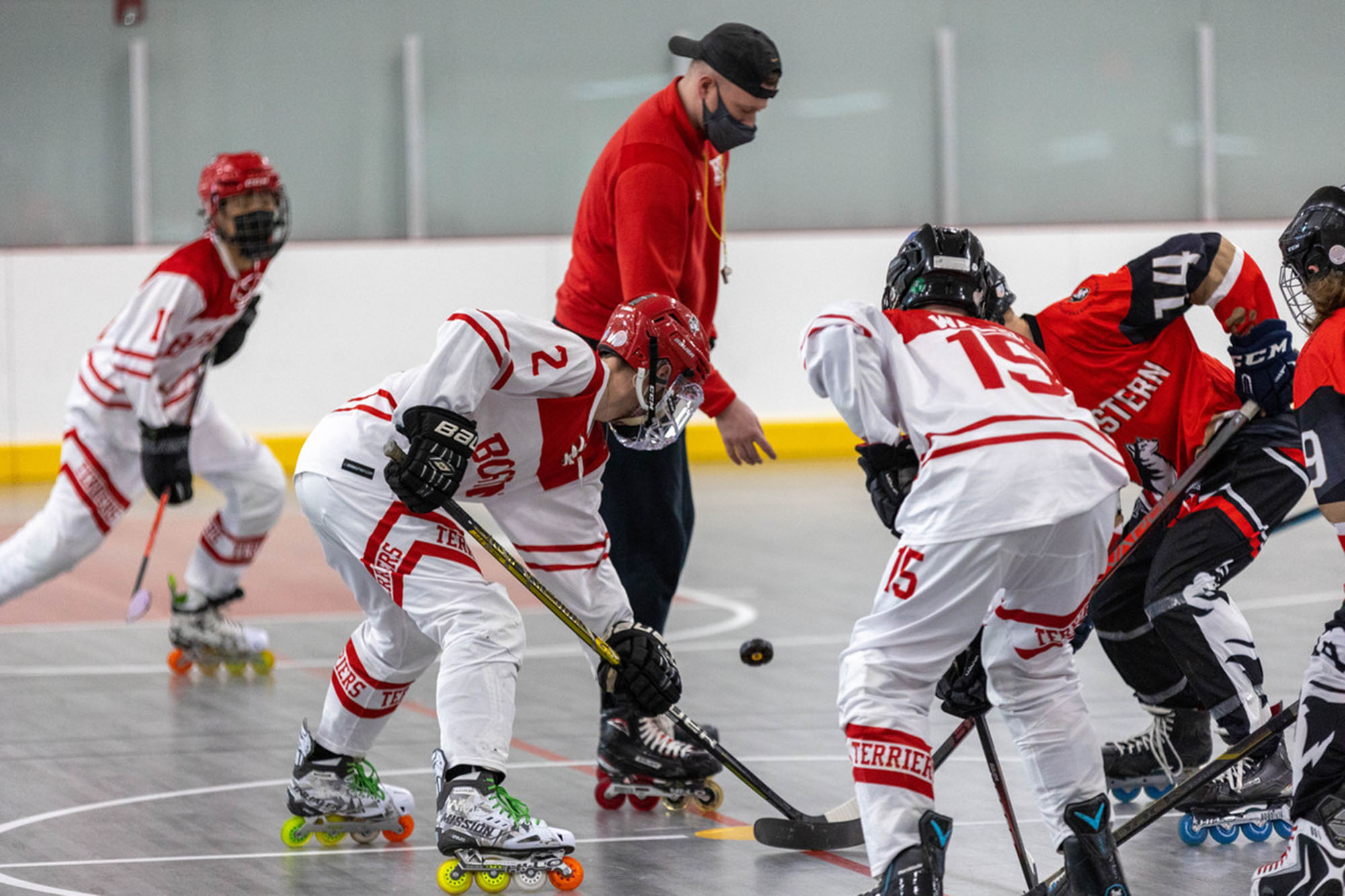 Photo of a faceoff. The BU Roller hockey team stands ready to one side as they face off against Northeastern. A ref is shown in red throwing a puck down as opposing players ready to get it.
