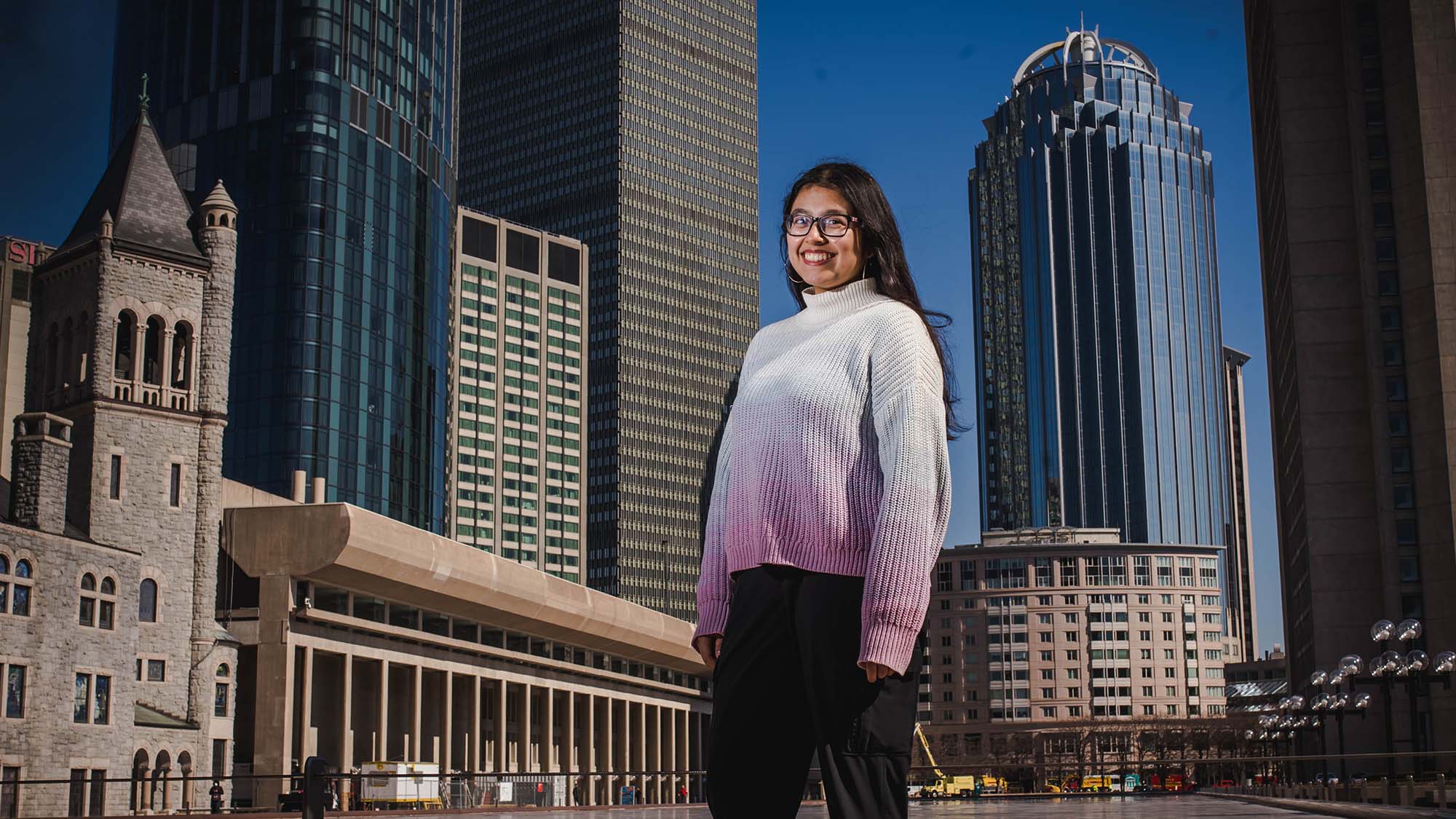 Photo of student Mira Dhakal, standing at the large reflection pool near the Christian Science Center. She smiles, wears a white and pink gradient sweater, and has long brown hair and glasses. The Boston city skyline and blue sky are seen behind her.