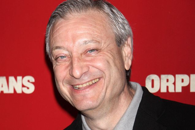 Candid photo of Fred Zollo as he stands in front of a red "Orphan" movie premiere background. An older white man with gray hair and wearing a grey shirt and black blazer smiles at the camera