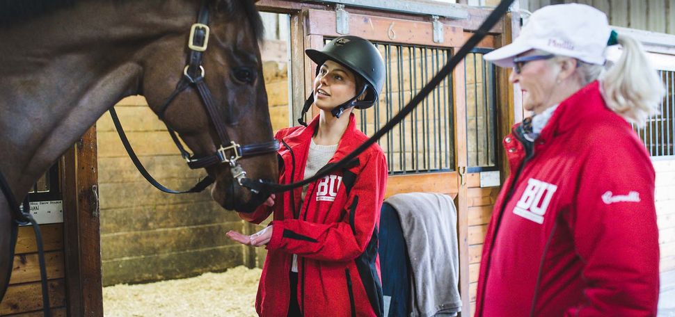 Photo of Lizzy Nevins, a young woman wearing a black riding helmet and red BU Equestrian jacket, standing next to BU Equestrian head coach Phyllis Cervelli, wearing a similar red jacket and white cap, in a barn barn. Lizzy is shown petting the head of a large dark brown horse with a black mane as Cervelli looks on to her right.