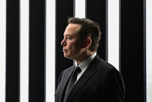 Photo of Elon Musk, a white man looking to the left as he wears a black suit ensemble and black tie. He stands in a dark room with lights going down in stripes behind him.