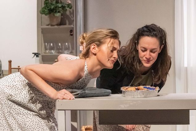 Photo of Katherine Schabe (CFA’22) (left) and Clara Francesca (right) performing at the Boston Playwrights’ Theatre in "Beasts". Two white women, one blonde and one brunette, lean over a table and sniff/inspect what looks to be a blueberry pie. Setting is a stage kitchen.