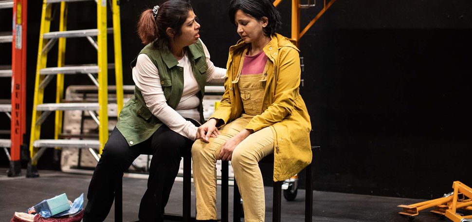 Photo of Mita Das, (left), a Bentley University Senior Lecturer of Mathematical Sciences and Prreeti Tiwari, right of South Windsor, CT, rehearsing for the Playwright's Theatre marathon. Das wears a green vest, white long sleeve, black pants and wears no shoes. She is a brown woman with brown hair. She sits on a stool and leans over to Tiwari, putting her hand on her knee. Tiwari, a brown woman with short black hair, looks forlorn, and wears bright yellow overalls.